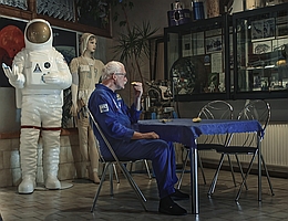 Man sieht den älteren Protagonisten in einem blauen Astronautenoverall an einem Tisch mit blauer Tischdecke sitzend ein Stück Obst essen. Der Boden ist gefliest, der Raum sieht nach Küche oder Esszimmer aus. Links im Hintergrund steht eine lebensgroße Figur mit einem angedeuteten Raumanzug, daneben eine lebensgroße Puppe mit wäremeregulierender Raumfahrerunterwäsche. Rechts sind in einer Vitrine bis zur Decke Raumfahrt-Devotionalien ausgestellt.