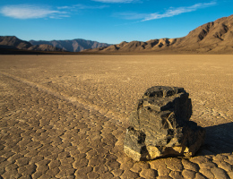 Blauer Himmel, ein dunkler Hang, davor eine beige flache Wüste: Darin liegt ein Stein, mit einer geraden Spur hinter sich.
