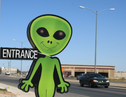 Eine lebensgroße Pappfigur eines stereotypischen grünen Außerirdischen an einer Straße, über die gerade ein Auto fährt.