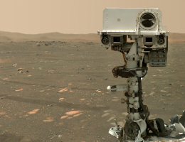 Perseverance steht in einer bräunlichen Ebene mit einigen flachen Steinen, am Horizont einige Hügel, der Rover „blickt“ zum Betrachter, neben ihm steht klein der Marshelikopter