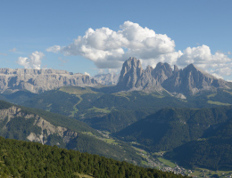 Ein Bergpanorama, in der Ferne schroff aufragende Dolomitenberge unter weißen Wölkchen, im Vordergrund Wälder und Wiesen Dolomitenlandschaft in Gröden: rechts die Langkofel-, links dahinter die Sellagruppe (Bild: CC-BY-SA 3.0 Wolfgang Moroder / Wikimedia Commons).