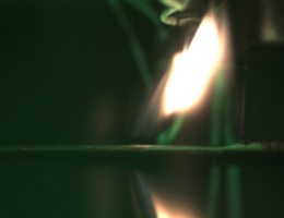 Seitenansicht der Verbrennung einer 2 mm dicken PMMA-Probe unter Mikrogravitation im Solid Combustion Experiment Module (SCEM) an Bord der ISS. Die Probe wurde am rechten Ende entzündet und die Flammen bewegen sich entgegen der erzwungenen Luftströmung auf der Probe fort. Die grüne Hintergrundbeleuchtung stammt von einer LED. Bild: JAXA